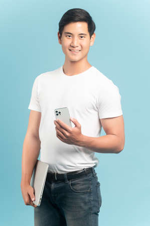 Nice asian guy in white t-shirt and jeans holding smartphone and laptop isolated on blue backgroundの写真素材