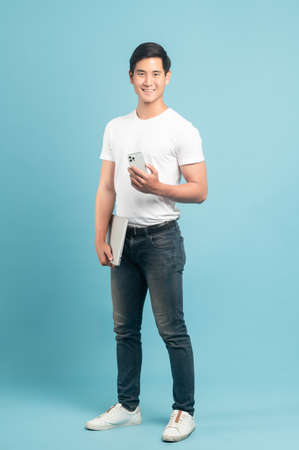 Nice asian guy in white t-shirt and jeans holding smartphone and laptop isolated on blue backgroundの写真素材