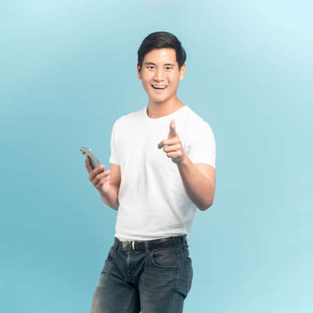 Nice asian guy in white t-shirt and jeans holding smartphone pointing at camera isolated on blue backgroundの写真素材