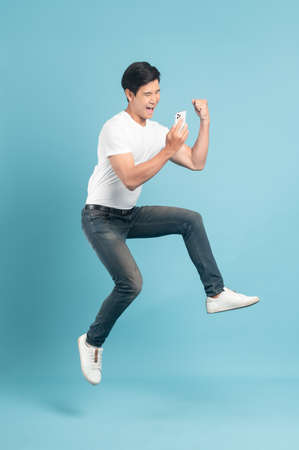 Jump shot of nice asian guy in white t-shirt and jeans holding smartphone isolated on blue backgroundの写真素材