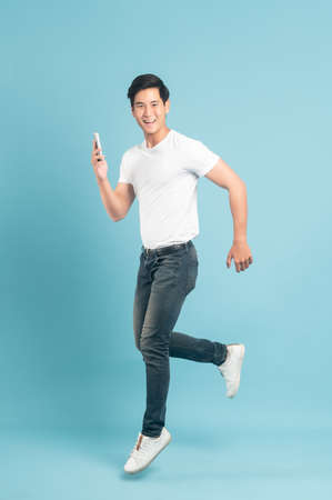 Jump shot of nice asian guy in white t-shirt and jeans holding smartphone isolated on blue backgroundの写真素材