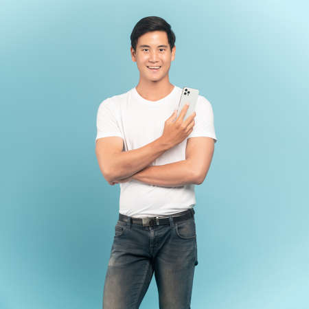 Nice asian guy in white t-shirt and jeans holding smartphone isolated on blue backgroundの写真素材