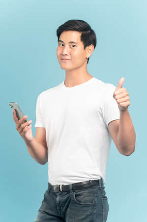 Nice asian guy in white t-shirt and jeans holding smartphone and show thumbs up isolated on blue backgroundの写真素材