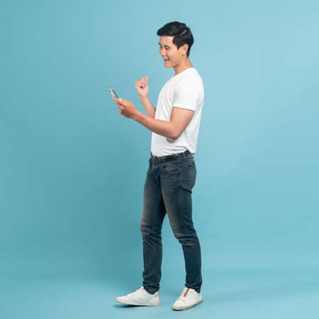Nice asian guy in white t-shirt and jeans holding smartphone isolated on blue backgroundの写真素材