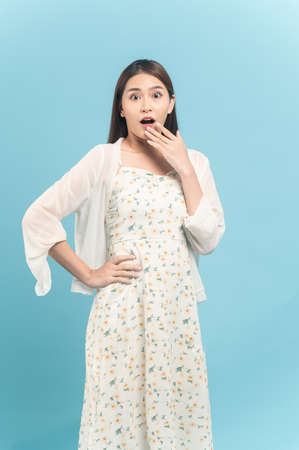 Beautiful young asian woman in white dress with flower pattern feeling surprise isolated on blue backgroundの写真素材