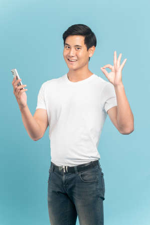 Nice asian guy in white t-shirt and jeans holding smartphone and make ok sign isolated on blue backgroundの写真素材