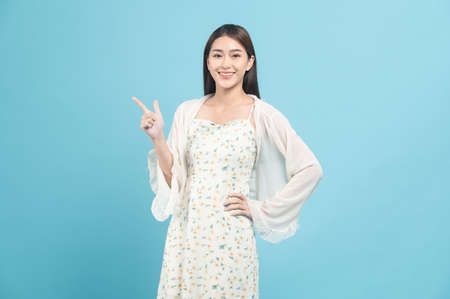Beautiful young asian woman in white dress with flower pattern pointing at copy space isolated on blue backgroundの写真素材