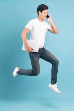 Nice asian guy in white t-shirt and jeans holding smartphone and laptop jumping isolated on blue backgroundの写真素材