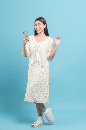 Beautiful young asian woman in white dress with flower pattern using smartphone isolated on blue backgroundの写真素材