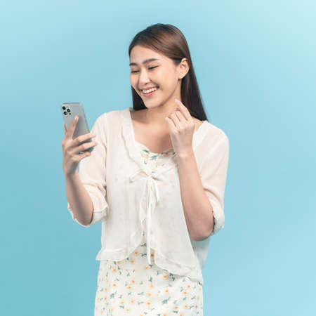 Beautiful young asian woman in white dress with flower pattern using smartphone isolated on blue backgroundの写真素材