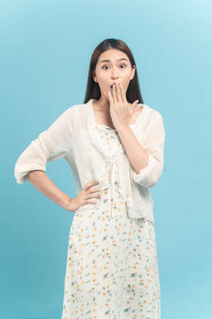 Beautiful young asian woman in white dress with flower pattern feeling surprise isolated on blue backgroundの写真素材