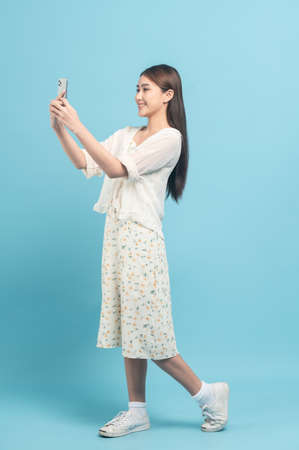 Beautiful young asian woman in white dress with flower pattern using smartphone isolated on blue backgroundの写真素材