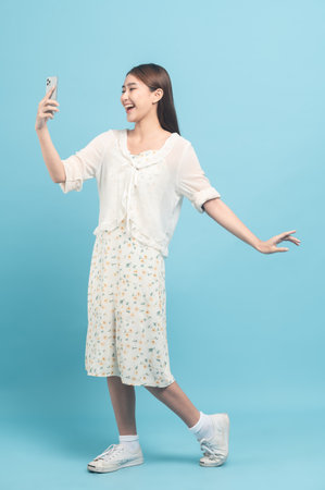 Beautiful young asian woman in white dress with flower pattern using smartphone isolated on blue backgroundの写真素材