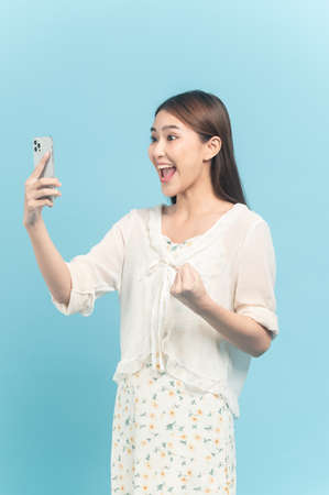 Beautiful young asian woman in white dress with flower pattern using smartphone feeling excite isolated on blue backgroundの写真素材