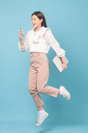 Young beautiful asian woman with smart casual cloth use smartphone and jump isolated on blue backgroundの写真素材