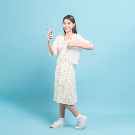 Beautiful young asian woman in white dress with flower pattern using smartphone show shumbs up isolated on blue backgroundの写真素材