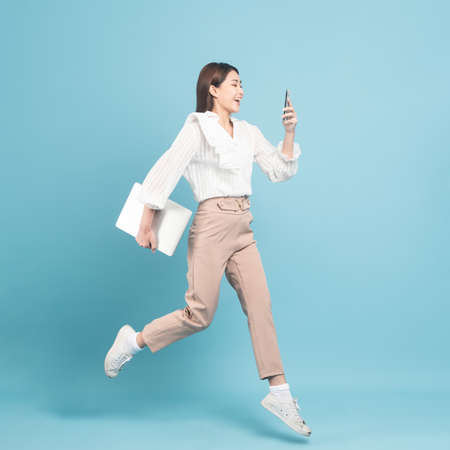 Young beautiful asian woman with smart casual cloth use smartphone and jump isolated on blue backgroundの写真素材