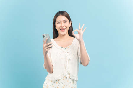 Beautiful young asian woman in white dress with flower pattern using smartphone show okay sign isolated on blue backgroundの写真素材