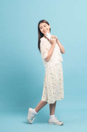 Beautiful young asian woman in white dress with flower pattern using smartphone isolated on blue backgroundの写真素材
