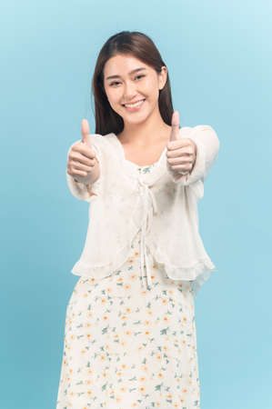 Beautiful young asian woman in white dress with flower pattern making thumbs up sign on both hands isolated on blue backgroundの写真素材