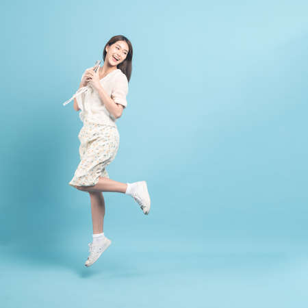 Beautiful young asian woman in white dress with flower pattern using smartphone and jump  isolated on blue backgroundの写真素材