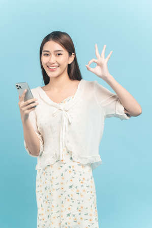 Beautiful young asian woman in white dress with flower pattern using smartphone show okay sign isolated on blue backgroundの写真素材