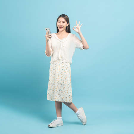 Beautiful young asian woman in white dress with flower pattern using smartphone show okay sign isolated on blue backgroundの写真素材