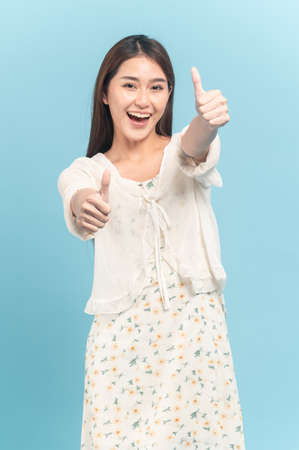 Beautiful young asian woman in white dress with flower pattern making thumbs up sign on both hands isolated on blue backgroundの写真素材