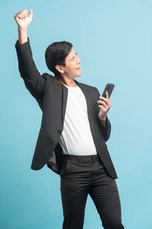 Good looking businessman portrait using smartphone with very excited emotion isolated on blue backgroundの写真素材