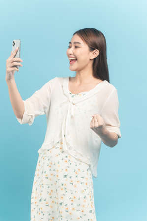 Beautiful young asian woman in white dress with flower pattern using smartphone feeling excite isolated on blue backgroundの写真素材