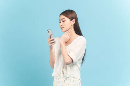 Beautiful young asian woman in white dress with flower pattern using smartphone examining isolated on blue backgroundの写真素材