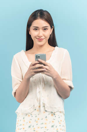 Beautiful young asian woman in white dress with flower pattern using smartphone isolated on blue backgroundの写真素材