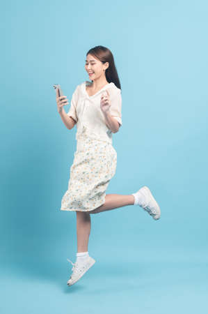 Beautiful young asian woman in white dress with flower pattern using smartphone and jump  isolated on blue backgroundの写真素材