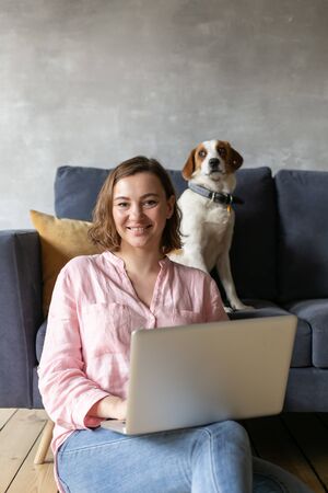A beautiful woman is working at her laptop at home with her dog.の写真素材