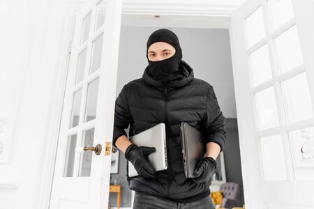 Thief looking at the camera with black balaclava stealing two modern expensive laptops. The burglar commits a crime in Luxury apartment with stucco.の写真素材