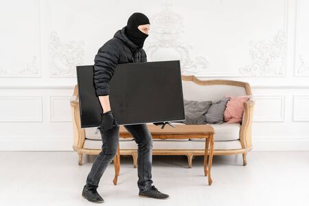 Thief with black balaclava stealing modern expensive television. Luxury apartment with stucco. Man burglar stealing tv set from houseの写真素材