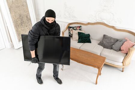The burglar commits a crime in Luxury apartment with stucco. Thief with black balaclava stealing modern expensive television.の写真素材