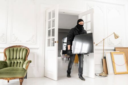 Thief with black balaclava stealing modern expensive television. Luxury apartment with stucco. Man burglar stealing tv set from houseの写真素材