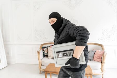 The burglar commits a crime in Luxury apartment with stucco. Thief with black balaclava stealing modern Electronic safe box.の写真素材