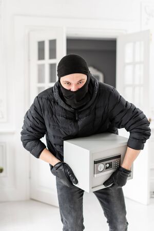 Thief looking at the camera with black balaclava stealing modern Electronic safe box. The burglar commits a crime in Luxury apartment with stucco.の写真素材