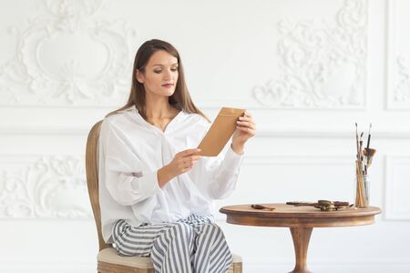A young beautiful girl in a luxurious interior sits on a chair, holds a letter in her hands and reads it. Soft focus.の写真素材