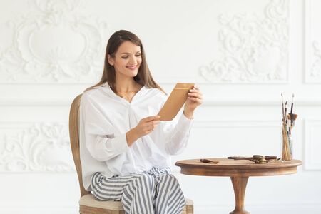 A young beautiful smailing girl in a luxurious interior sits on a chair holding a love letter from her boyfriend and reads it. Romantic mood on Valentines Day. Soft focus.の写真素材