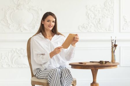 A young beautiful girl in a luxurious interior sits on a chair, holds a letter in her hands and reads it. Soft focus.の写真素材