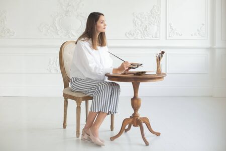 An inspired girl artist holds a palette and a brush in her hands, sits on a chair in the interior of the workshop and paints a picture at the table. Soft focus.の写真素材