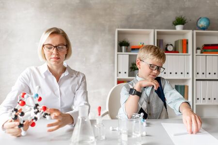 Blond schoolboy write properties of chemical element into his notebook under guidance of his charming teacherの写真素材