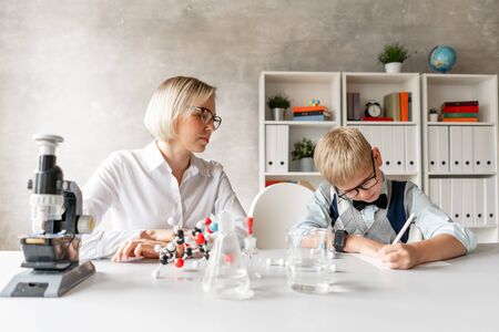 Blond schoolboy write properties of chemical element into his notebook under guidance of his charming teacherの写真素材