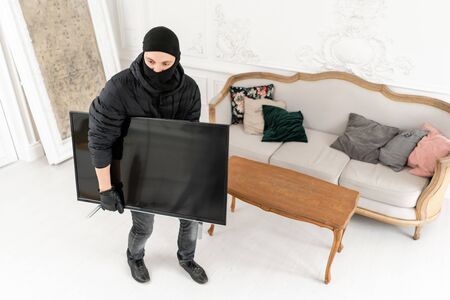 Thief with black balaclava stealing modern expensive television. Luxury apartment with stucco. Man burglar stealing tv set from houseの写真素材