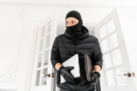Thief with black balaclava stealing two modern expensive laptops. The burglar commits a crime in Luxury apartment with stucco.の写真素材