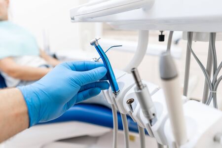Close-up hand of dentist in the glove holds dental high speed turbine. The patient in blue chair at the background. Office where dentist conducts inspection and concludes.の写真素材