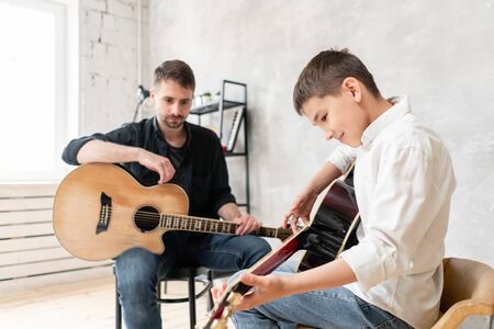 Two brothers learn to play acoustic guitar. The older brother tunes the guitar while the younger one runs his finger along the stringsの写真素材